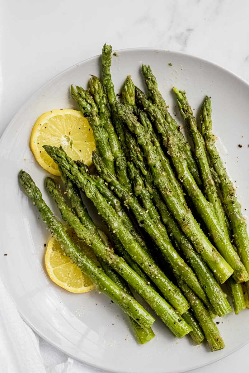 Close view of seasoned, plump asparagus on a round white plate with two lemon wheels.