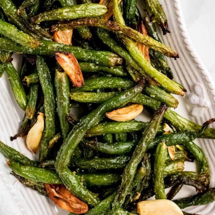 Close view of a white serving platter piled high with seasoned grilled green beans and garlic cloves.