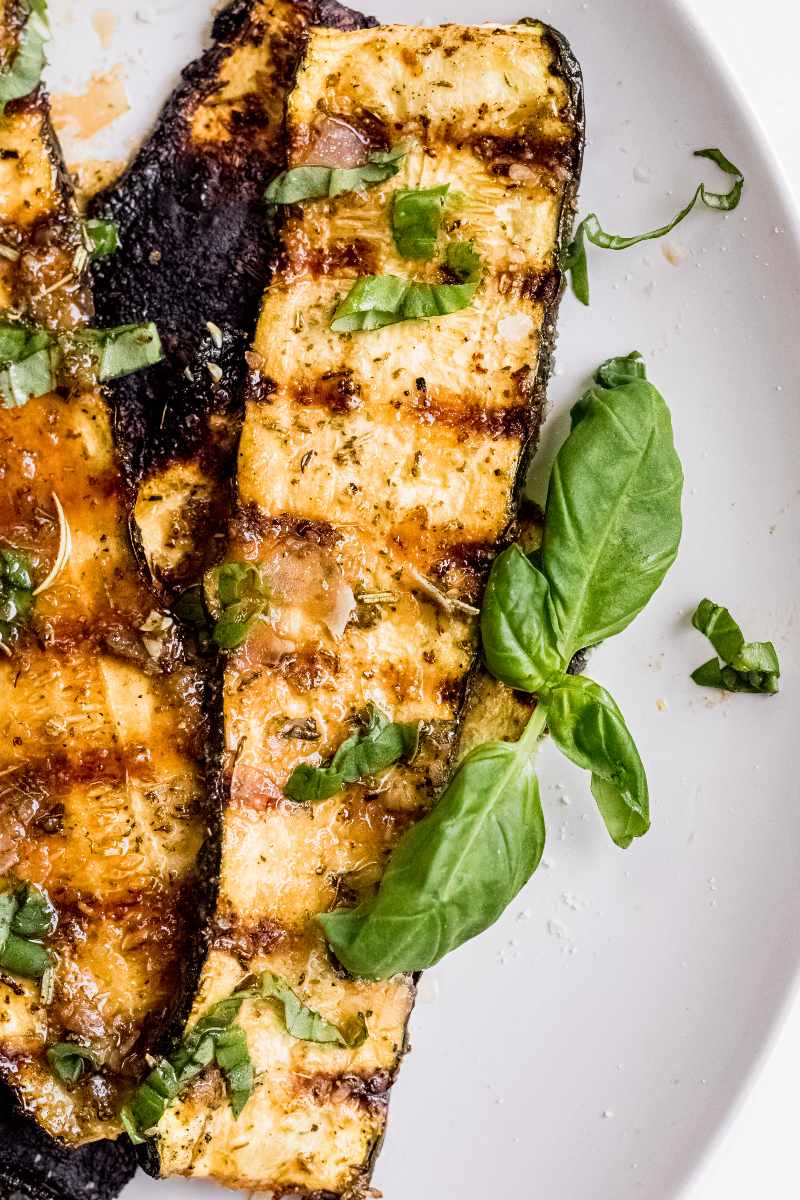 Close view of a tender grilled zucchini plank on a plate with fresh basil leaves beside it.