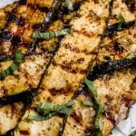 Grilled parmesan zucchini slices piled on parchment paper garnished with ribbons of fresh basil.