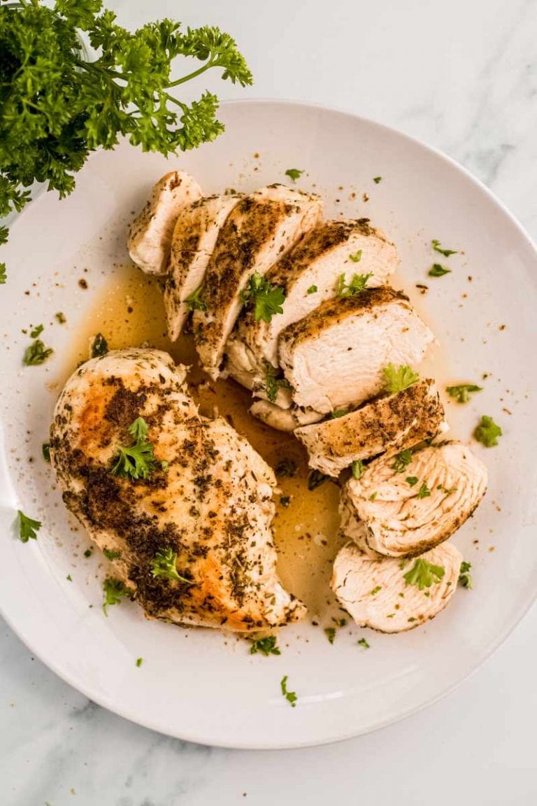 Two cooked chicken breasts on a white plate with flavorful juices, one whole and one sliced.