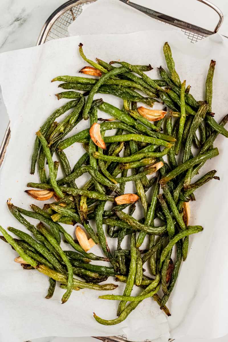 Overhead of grill basket lined with parchment paper with finished grilled green beans and garlic cloves on top.