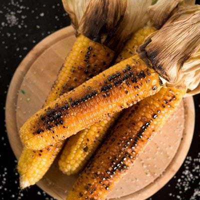 Close view of four grilled corn on the cob piled on a round wooden cutting board.