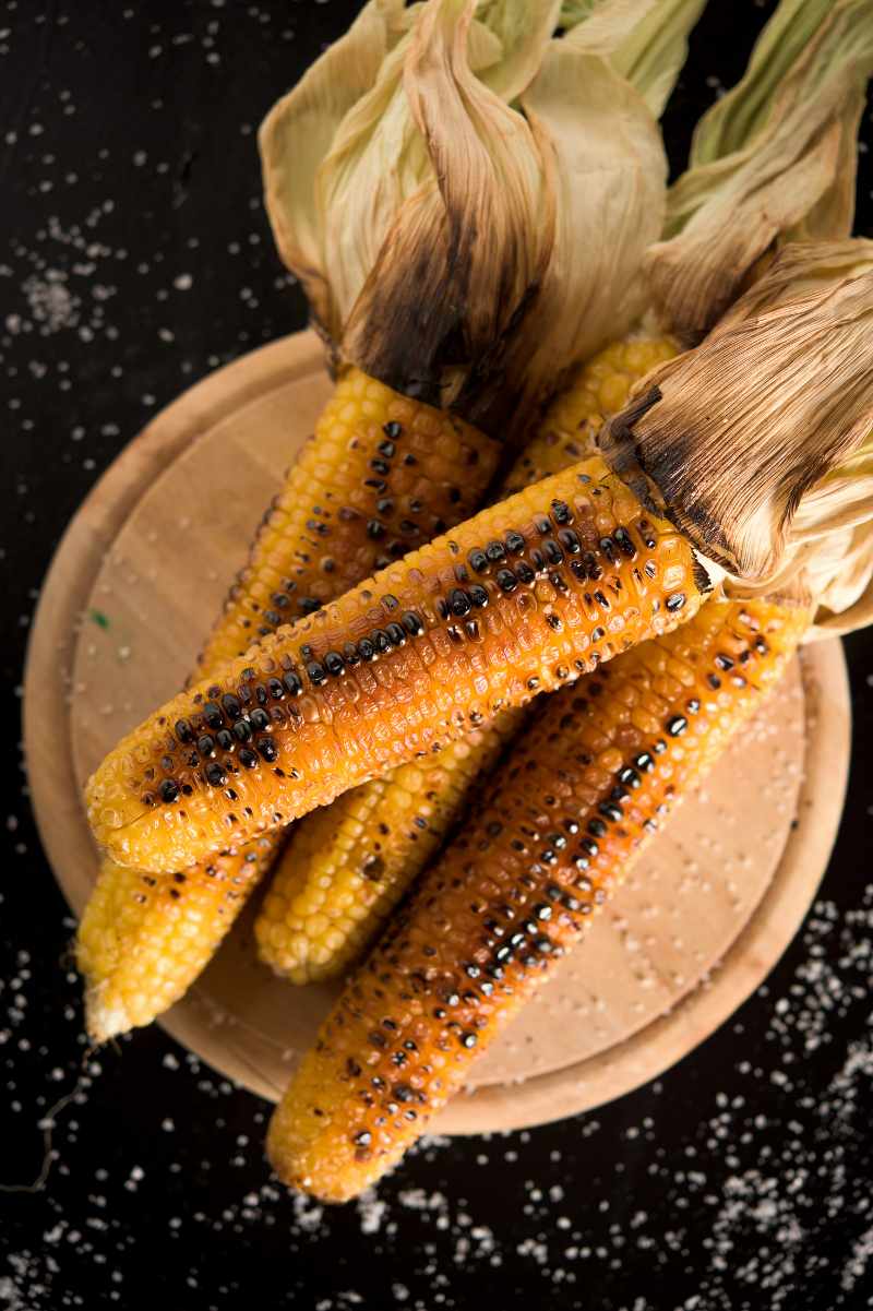 Close view of four grilled corn on the cob piled on a round wooden cutting board.