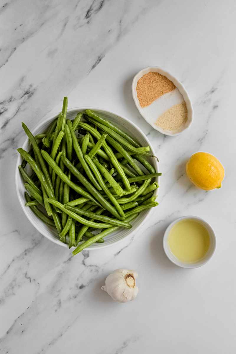 Overhead of ingredients needed to make grilled garlic green beans, including fresh green beans, a head of garlic, and lemon.