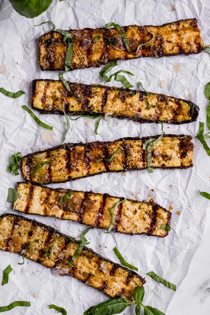 Birdseye of individual slices of grilled zucchini arranged from top to bottom on parchment paper scattered with fresh basil ribbons.