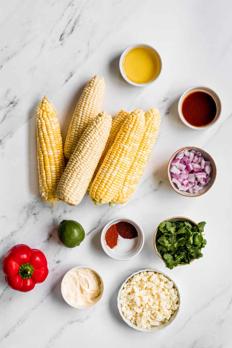 Overhead of ingredients needed to make mexican corn salad, including fresh corn, red bell pepper, lime, and seasonings.