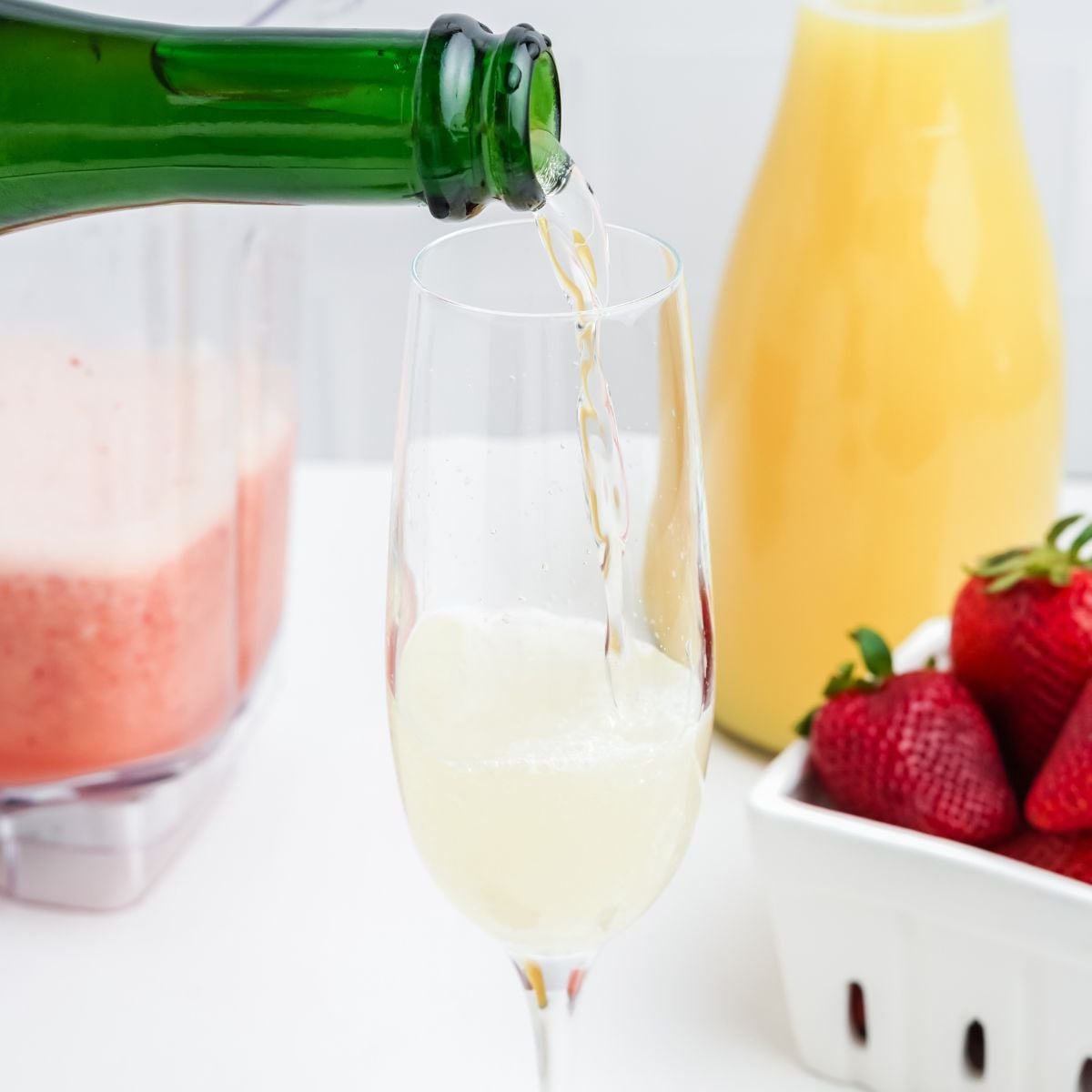 Sparkling wine being poured into a champagne flute, with strawberry puree and orange juice in the background