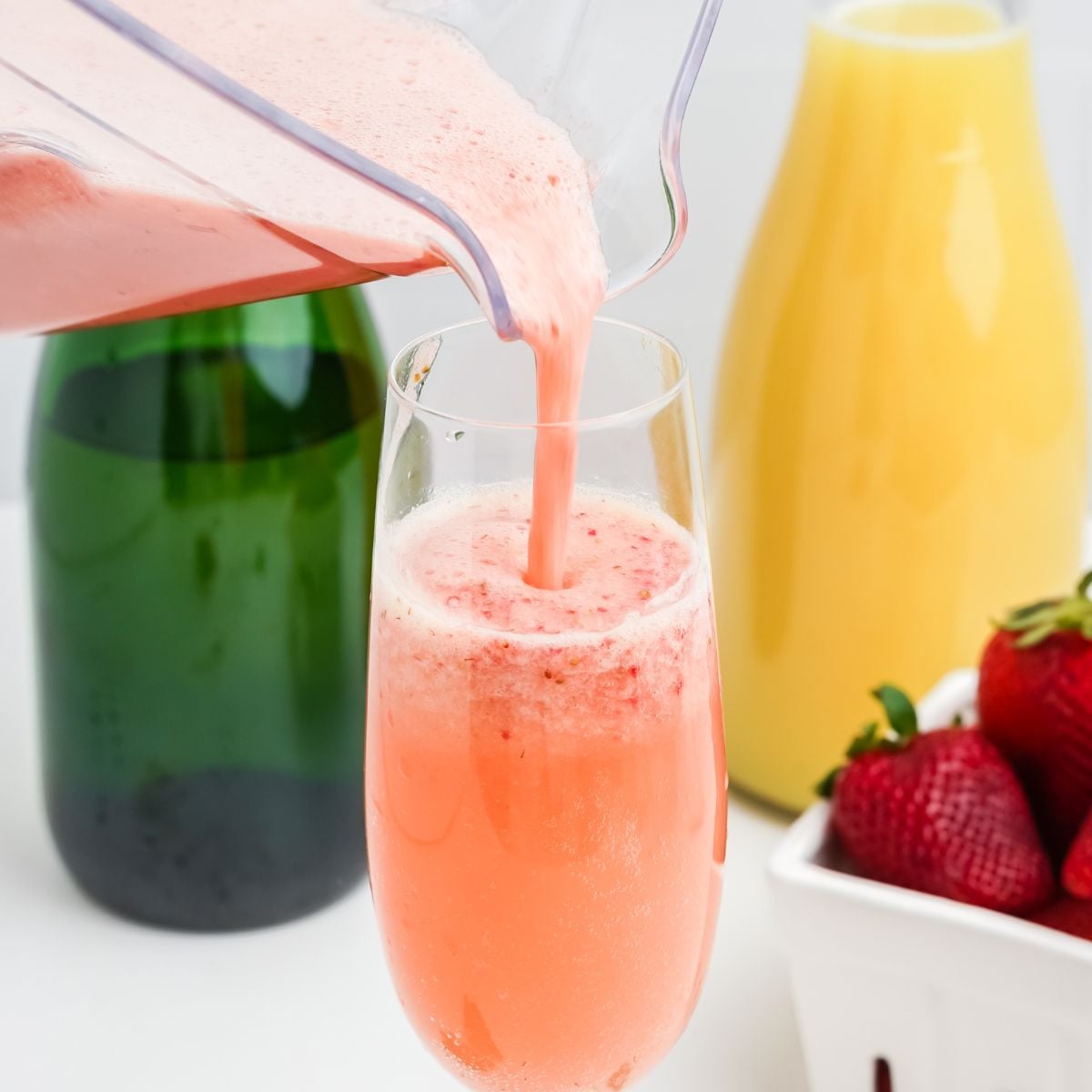 Strawberry puree being poured from a blender into a champagne flute next to sparkling wine and orange juice
