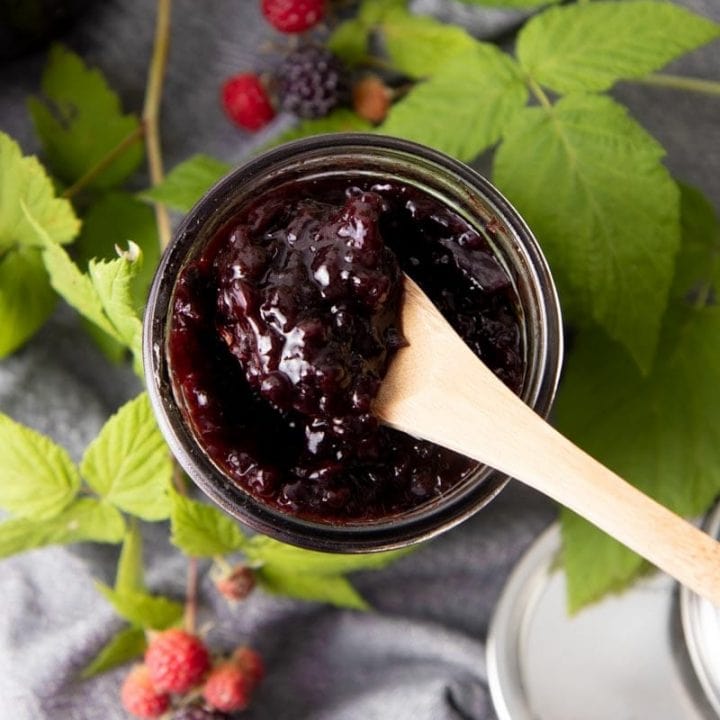 Birdseye view of a wooden spoon scooping dark, fruity jam from an open jar.