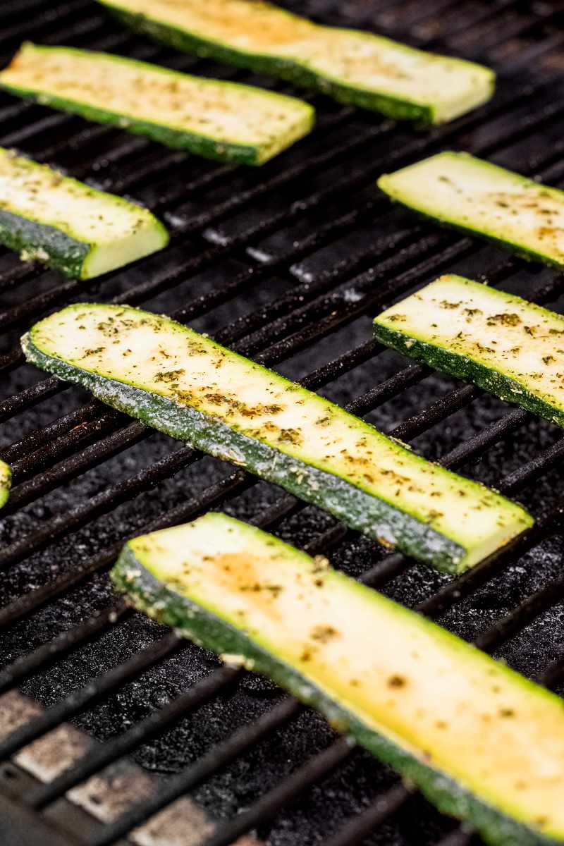 Seasoned raw zucchini slices on grill grates beginning to cook.