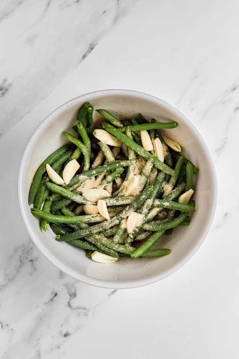 Overhead of blanched green beans in a large white bowl with garlic cloves and seasoning over top.