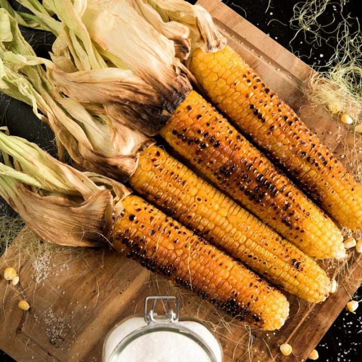 Top view of grilled corn with charred kernels on a wooden serving board, a container of salt nearby.