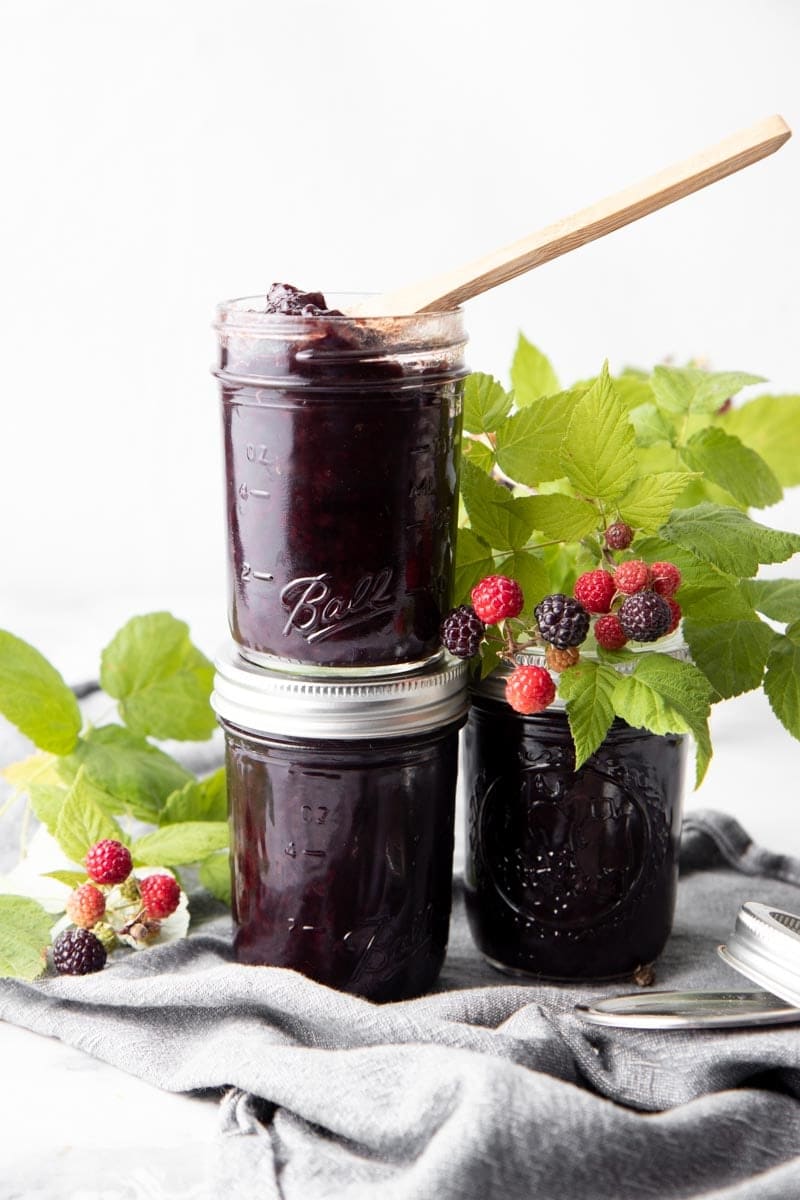A trio of half-pint jars of black raspberry jam, two stacked on top of one another with fresh raspberries and leaves around them.