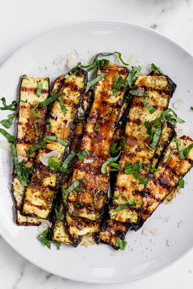 Tender grilled zucchini piled onto a white plate and garnished with grated parmesan cheese and ribbons of fresh basil.