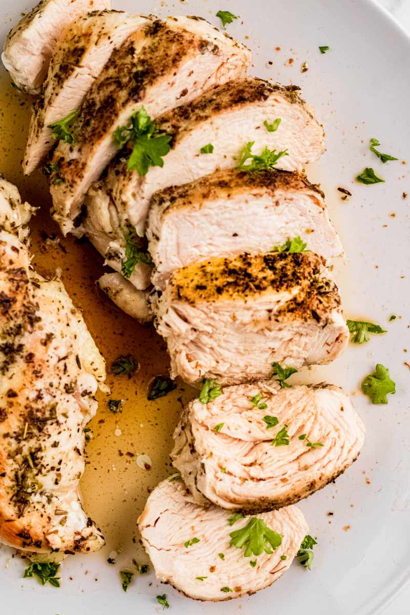 Juicy slices of chicken arranged on a white plate in flavorful chicken juices and garnished with chopped herbs.