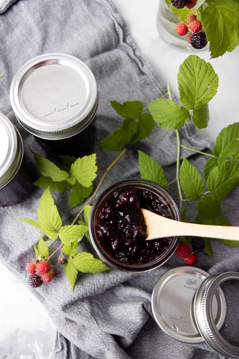 Top view of a wooden spoon digging into the black raspberry jam in an open half-pint jar.