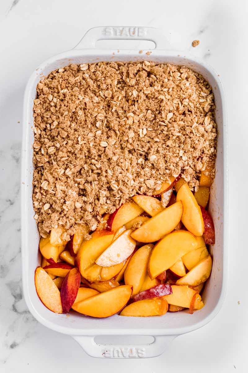 A peach crisp is being assembled in a rectangular baking dish. Only half the topping is on so far.