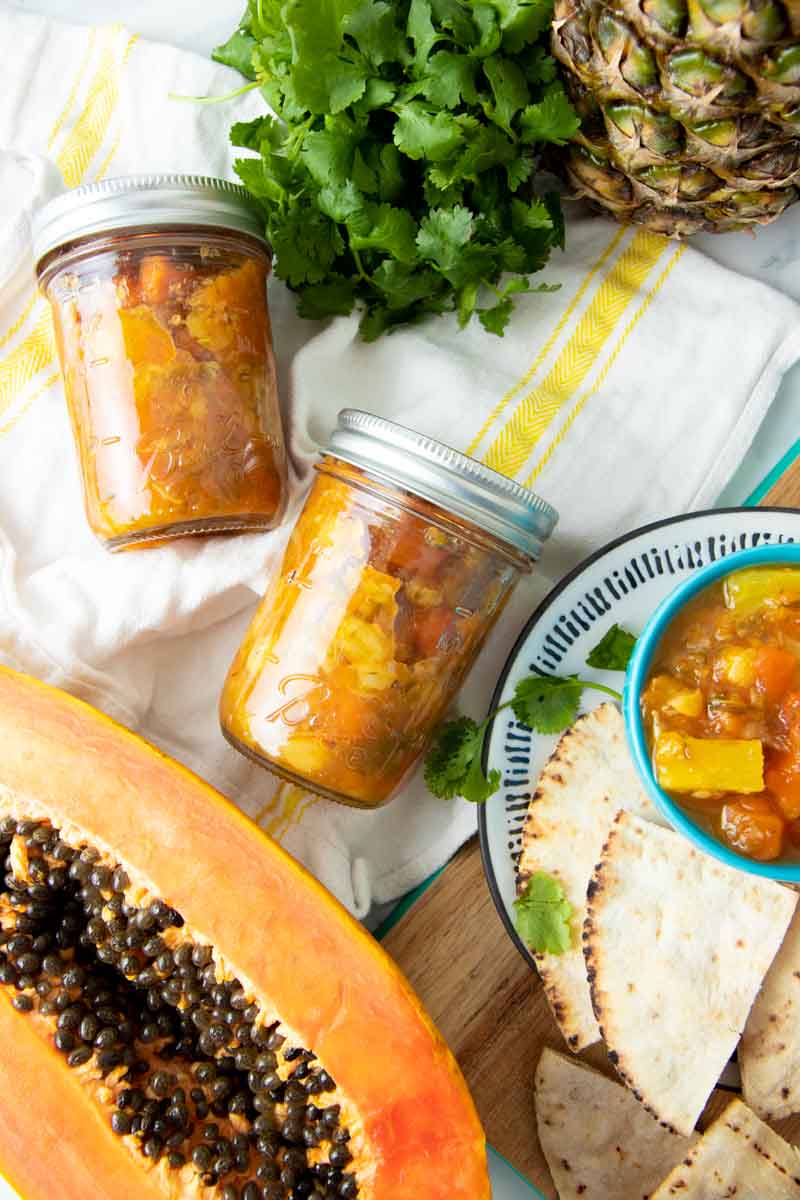 Two sealed jars of pineapple salsa lay on a folded towel between a halved papaya and a bunch of cilantro.