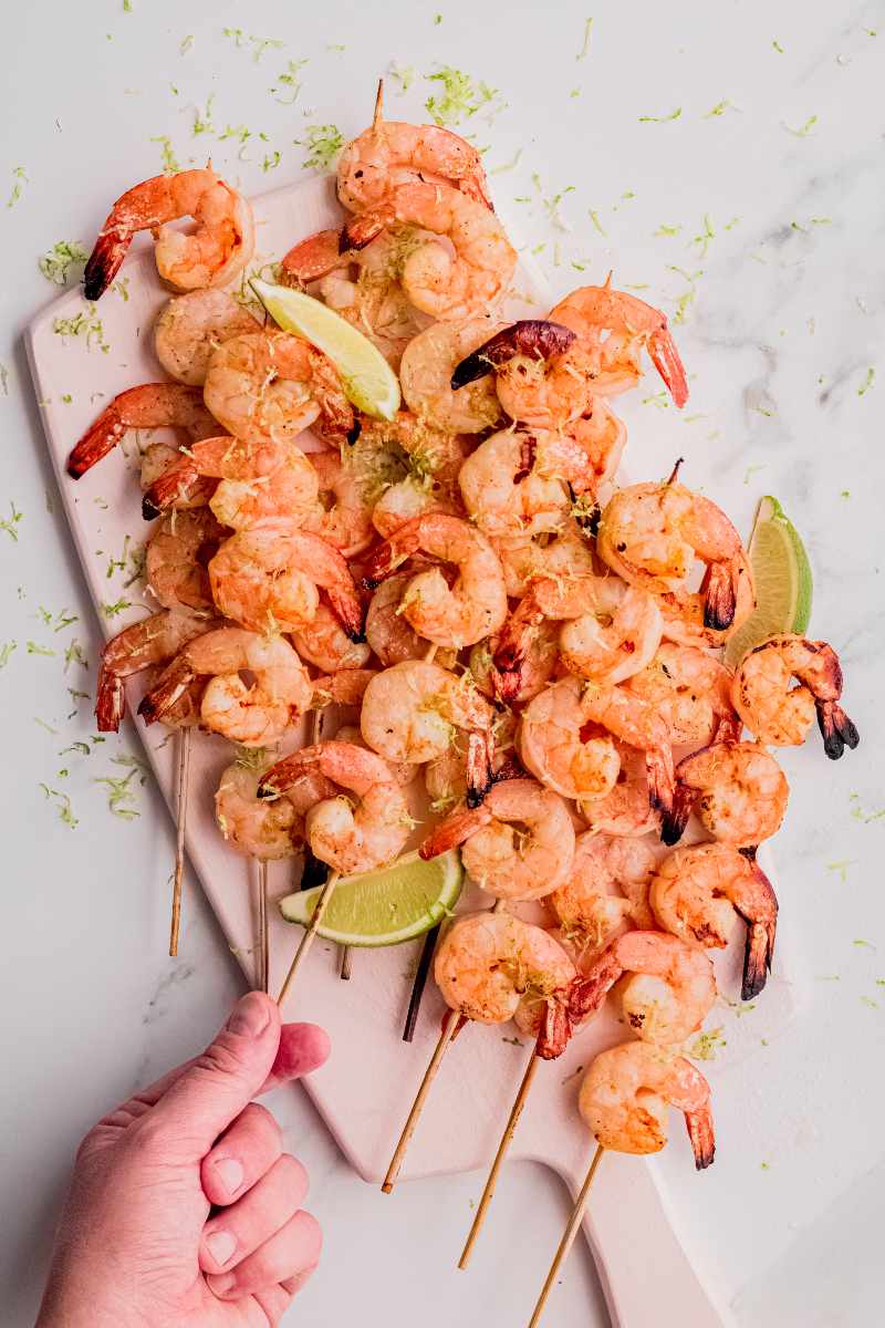 A hand grabs a grilled shrimp kabob from a serving board.