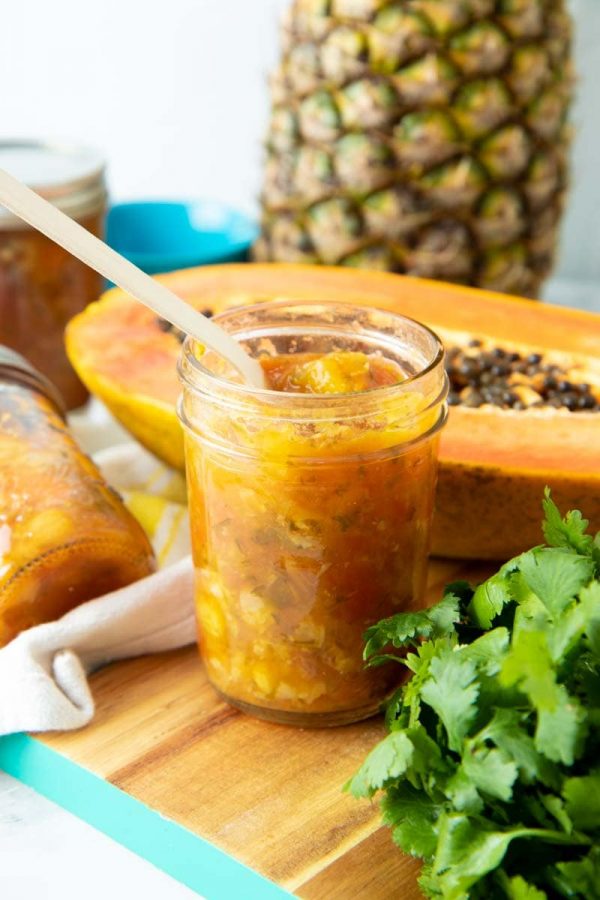 A spoon sticks out of an open jar of pineapple salsa. A halved papaya and a whole pineapple are in the background