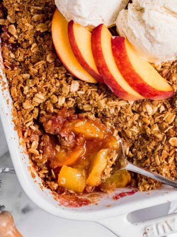 Overhead of a spoon dipping into a peach crisp. Fresh peach slices are arranged on the top of the middle of the crisp.