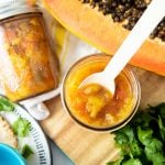A white spoon dips into a jar of pineapple salsa. A second jar, halved papaya, and loose cilantro surround the jar
