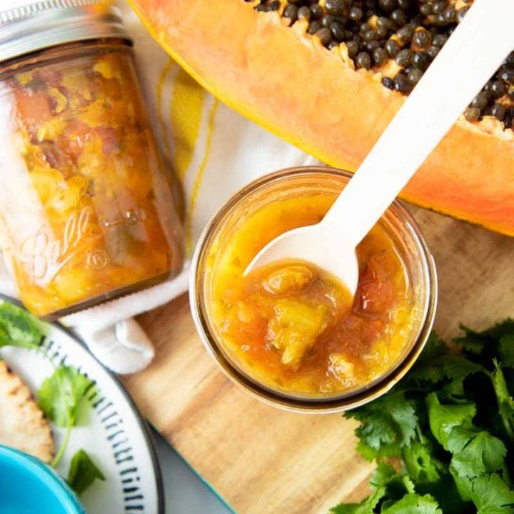 A white spoon dips into a jar of pineapple salsa. A second jar, halved papaya, and loose cilantro surround the jar