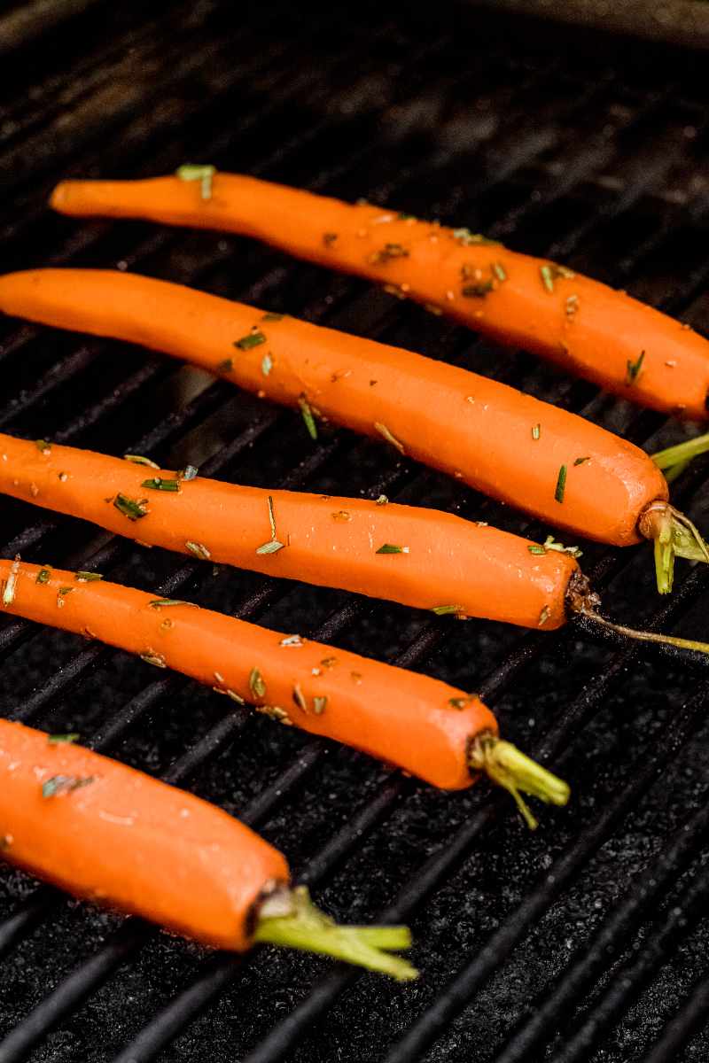 Seasoned carrots on gas grill grates with fresh herbs and seasonings.