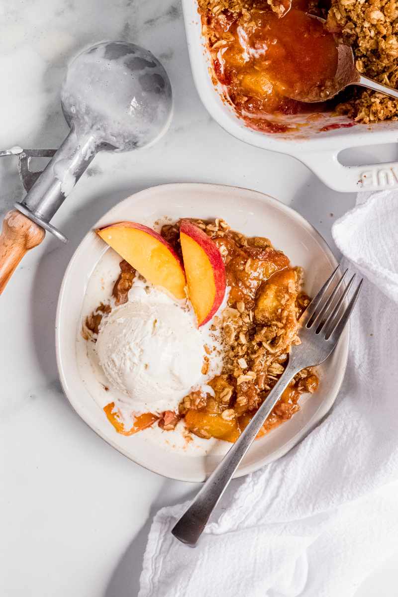 A bowl of peach crisp with a scoop of vanilla ice cream. A fork rests on the side of the dis