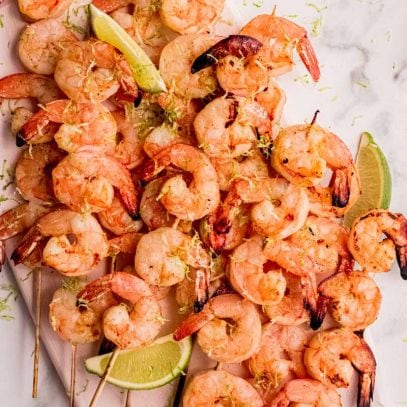 A white rectangular serving platter loaded with shrimp kabobs with lime wedges and lime zest garnish.