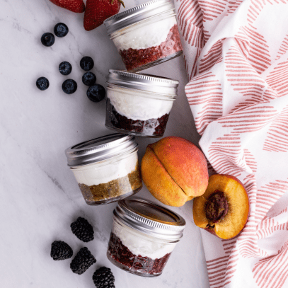 Four jars of yogurt with fruit on the bottom lay on their sides, surrounded by fresh fruit and a tea towel.