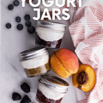 Four jars of yogurt with fruit on the bottom lay on their sides, surrounded by fresh fruit and a tea towel. A text overlay reads "fruit on the bottom yogurt jars"