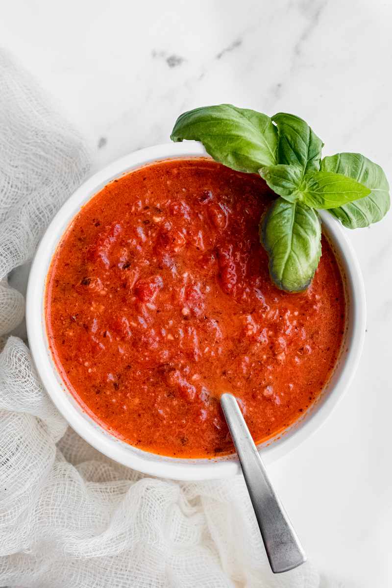 A white bowl of roasted red pepper sauce with a spoon in it and a fresh basil sprig tucked beside it.