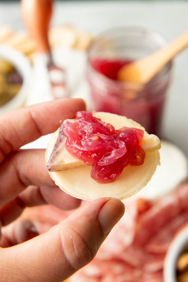 Close view of a hand holding a cracker with a slice of cheese and red preserves on top.