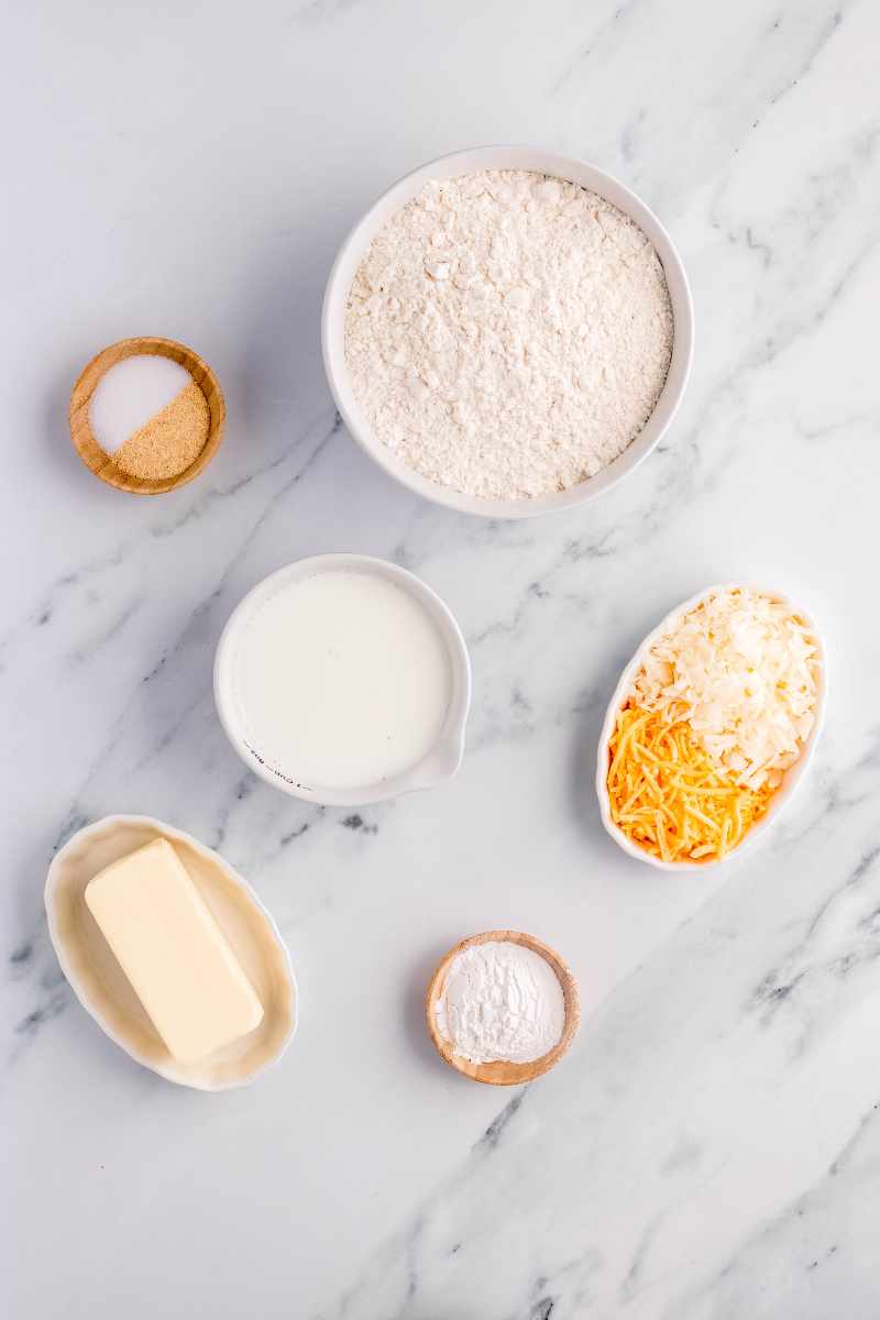 Overhead of ingredients, including butter, flour, and milk, in bowls on a marble counter.