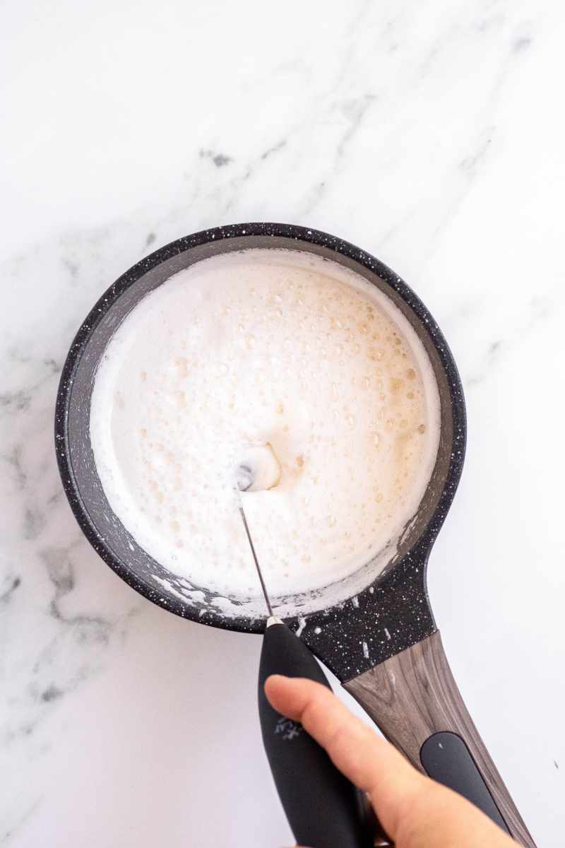 A hand holds a milk frother into a small saucepan of steamed milk.
