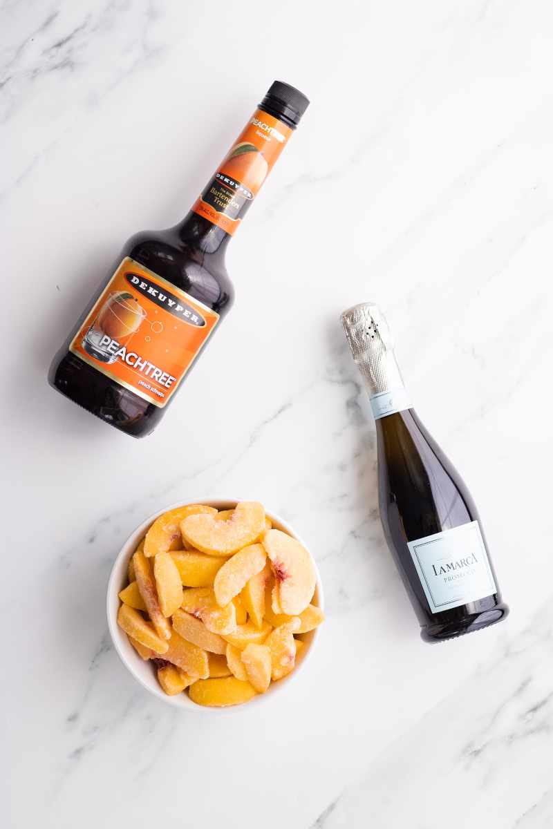 Three ingredients needed to make peach bellini cocktails including frozen peach slices, peach schnapps, and prosecco.