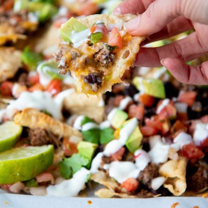 A hand holds up a nacho chip loaded with toppings including seasoned ground beef, black beans, and avocado.