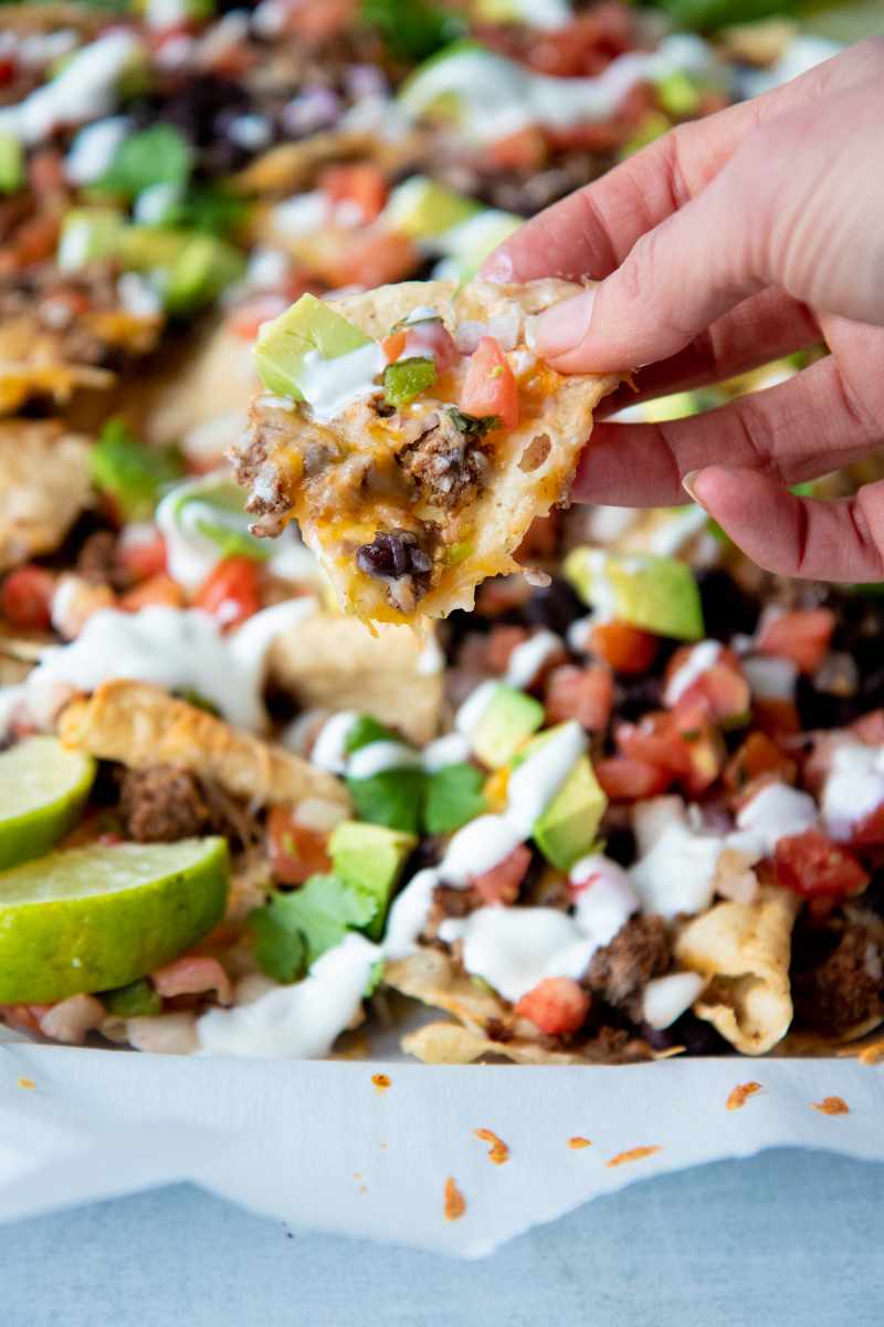 A hand holds up a nacho chip loaded with toppings including seasoned ground beef, black beans, and avocado.