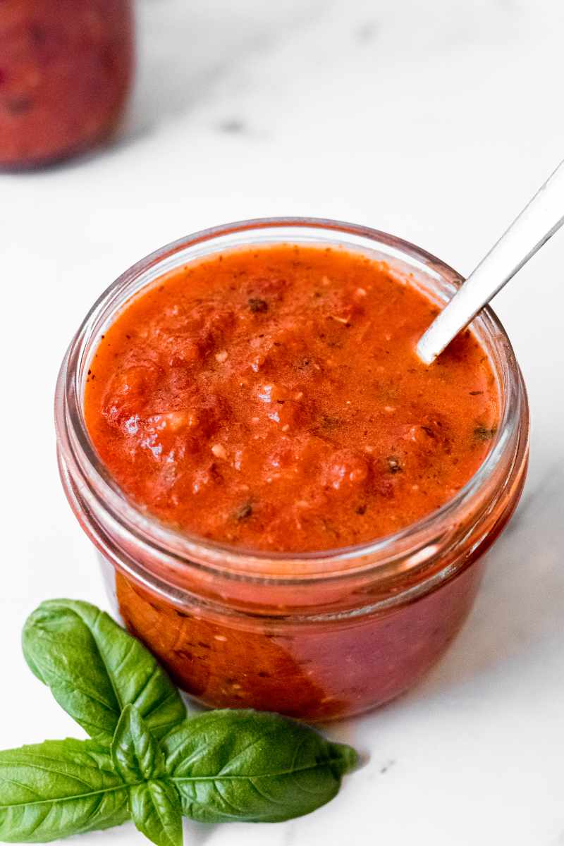 A spoon rests in a small glass jar of homemade red pepper sauce, a sprig of fresh basil beside it.