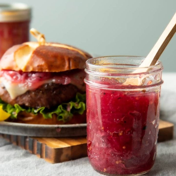 A jar of pink-hued red onion and port jam with a wooden spoon in it stands next to a plate with a cheeseburger topped with onion jam.
