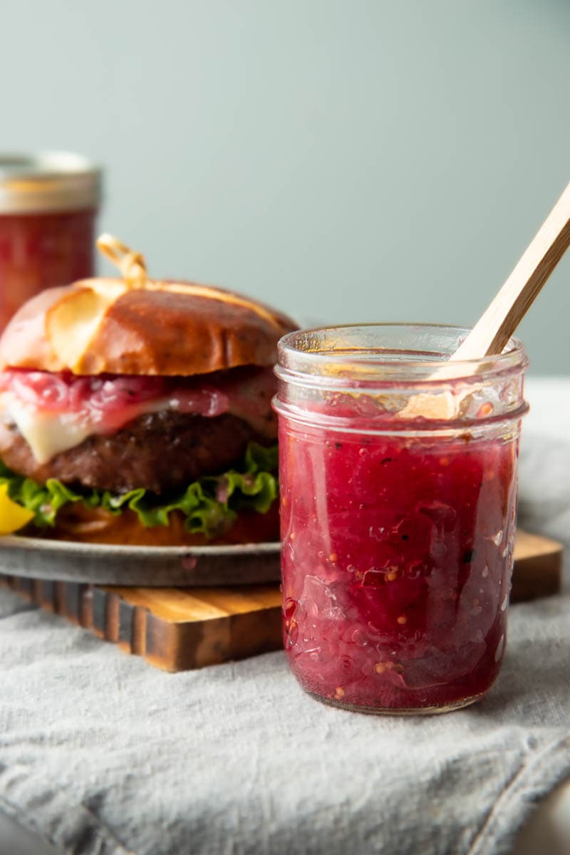 A jar of pink-hued red onion and port jam with a wooden spoon in it stands next to a plate with a cheeseburger.