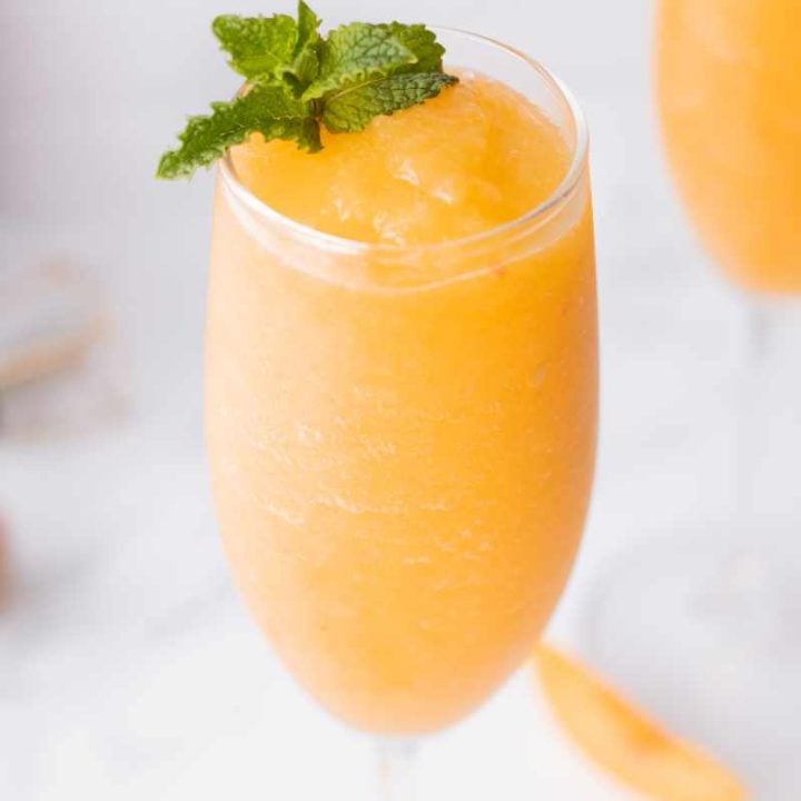 Close view of a blended peach cocktail in a champagne glass garnished with a fresh mint sprig.