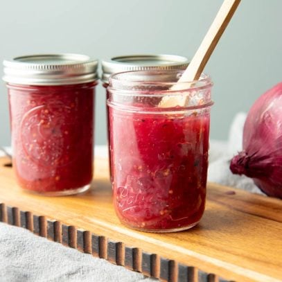 Three jars of onion jam stand on a wooden board, the jar in front is open and has a wooden spoon in it.