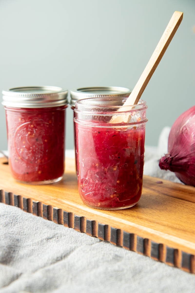 Three jars of onion jam stand on a wooden board, the jar in front is open and has a wooden spoon in it.