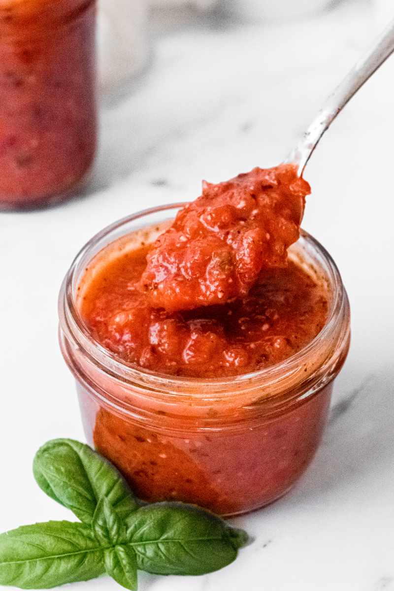 A spoon scoops red pepper sauce from a small glass jar.