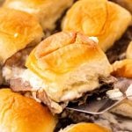 A metal spatula lifts a baked roast beef slider from the center of a pan of sandwiches.