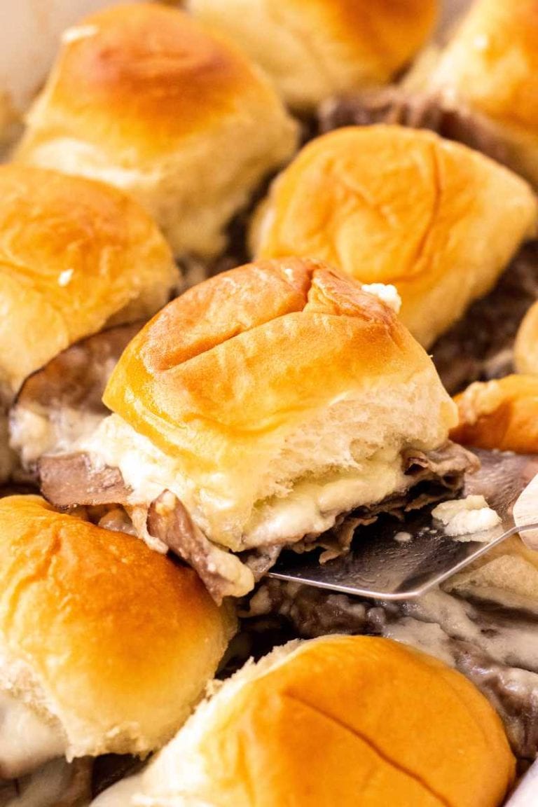 A metal spatula lifts a baked roast beef slider from the center of a pan of sandwiches.
