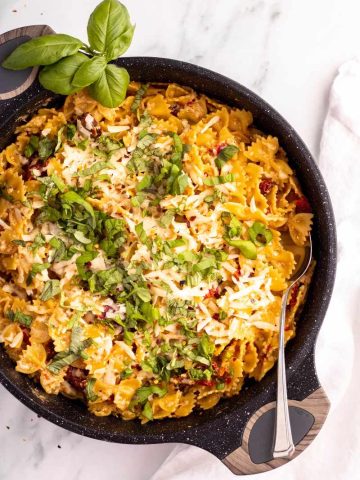 A large serving spoon is tucked into a saucepan of creamy sun-dried tomato pasta, a sprig of fresh basil alongside.